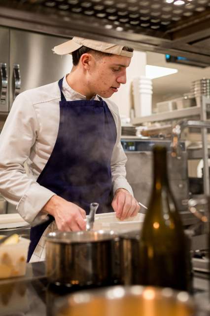 Jeune cuisinier du restaurant La Table d’Elka en pleine préparation en cuisine, portant un tablier bleu et une tenue de chef blanche.