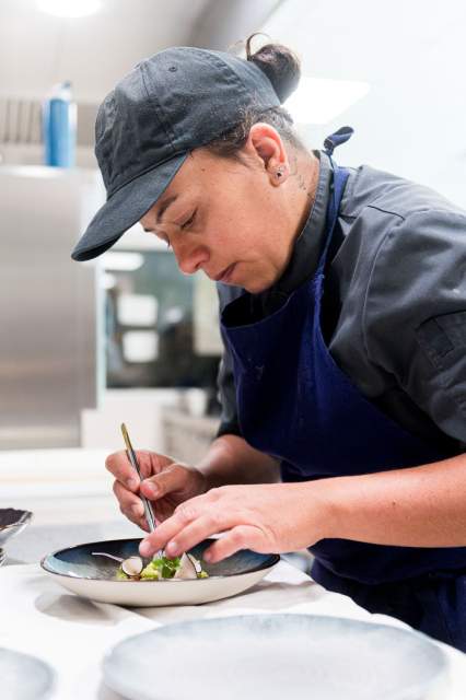 Cheffe du restaurant La Table d’Elka concentrée sur le dressage d’une assiette gastronomique en cuisine, portant un tablier bleu et une casquette noire.