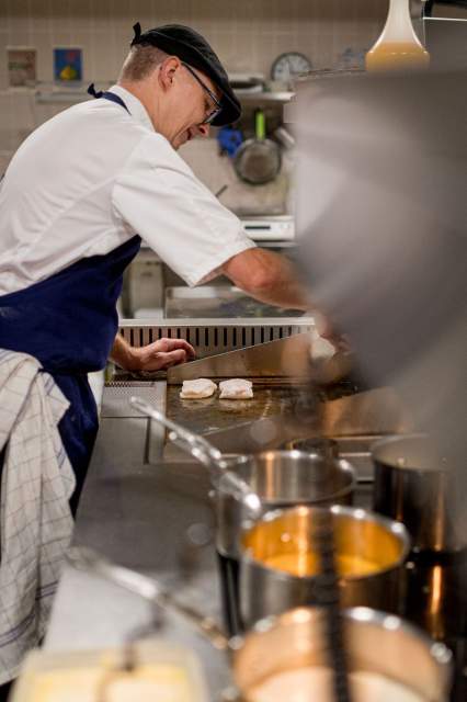 Chef du restaurant La Table d’Elka cuisinant des poissons à la plancha dans la cuisine, avec des casseroles en premier plan.