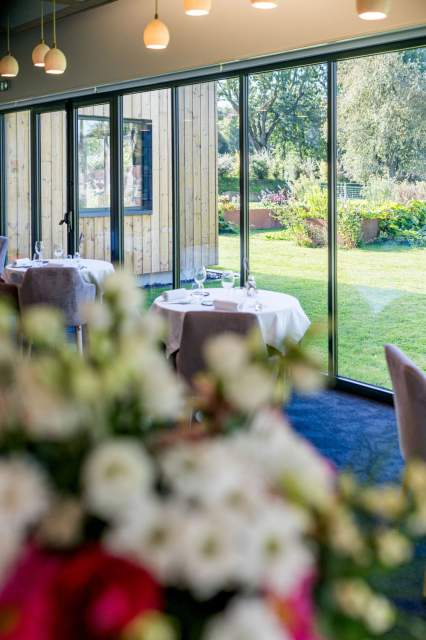Salle du restaurant La Table d’Elka avec vue sur le jardin, tables dressées et bouquet de fleurs au premier plan.