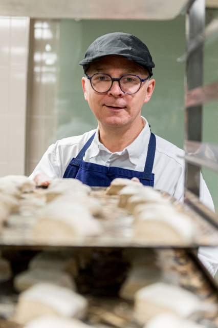 Chef du restaurant La Table d’Elka en cuisine, portant un tablier bleu et une casquette noire, devant une plaque de préparation culinaire.