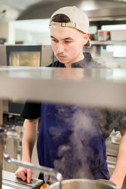 Jeune cuisinier du restaurant La Table d’Elka concentré sur la préparation d’un plat chaud en cuisine, entouré de vapeur.