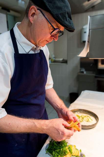 Chef du restaurant La Table d’Elka concentré sur le dressage d’une assiette en cuisine, portant un tablier bleu et une casquette noire.