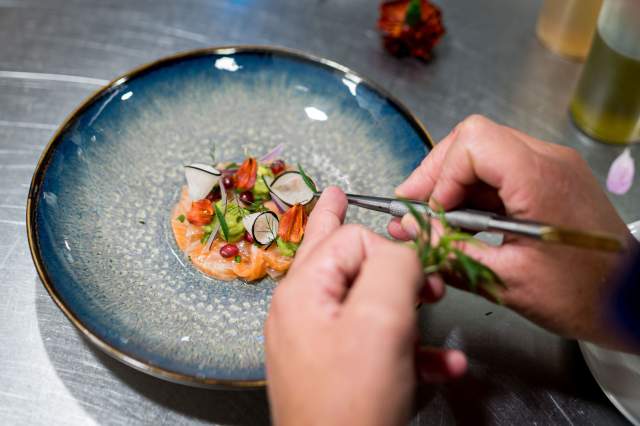 Dressage précis d’une entrée au restaurant La Table d’Elka, un chef disposant des herbes fraîches sur un plat coloré avec des légumes et du poisson.