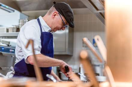 Chef du restaurant La Table d’Elka en train de cuisiner dans la cuisine professionnelle, portant un tablier bleu et une casquette noire.