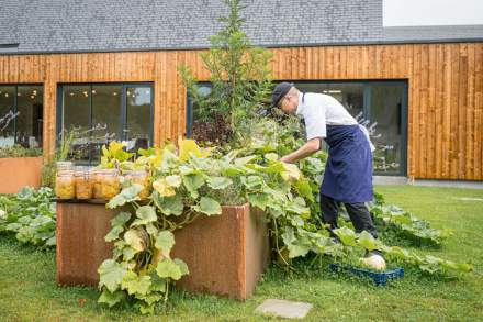 Chef du restaurant La Table d’Elka récoltant des légumes dans le potager du restaurant, devant la façade en bois du bâtiment.