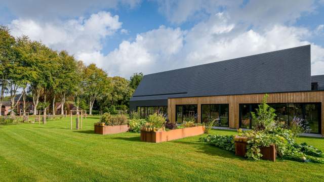 Extérieur du restaurant La Table d’Elka avec façade en bois, potager et pelouse verdoyante sous un ciel bleu partiellement nuageux.