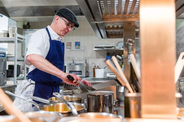 Chef du restaurant La Table d’Elka en train de cuisiner dans la cuisine professionnelle, portant un tablier bleu et une casquette noire.