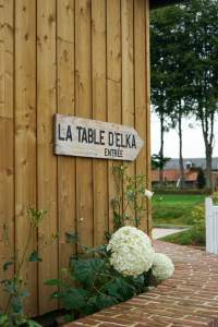 Panneau en bois indiquant l’entrée du restaurant La Table d’Elka, fixé sur la façade extérieure entourée de fleurs et de verdure.