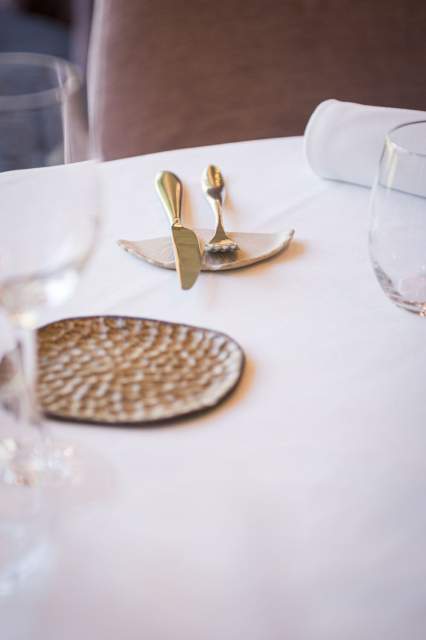 Détail d’une table dressée au restaurant La Table d’Elka, avec set de table tressé, couverts et verres disposés sur une nappe blanche.