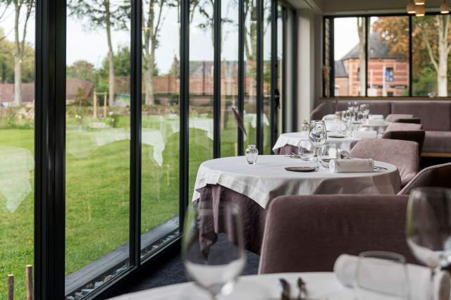 Tables du restaurant La Table d’Elka dressées le long des baies vitrées avec vue sur le jardin et les bâtiments environnants.